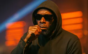 A man in a dark hoodie and sunglasses holds a microphone to his lips, performing under stage lights.