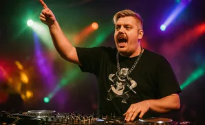 A DJ with blonde hair and a mustache, wearing a black t-shirt, points up with his right hand while playing on a DJ console.