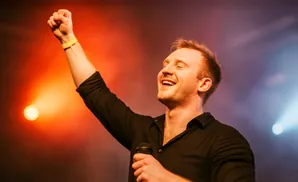 A man sings into a microphone with his fist raised on a brightly lit stage.