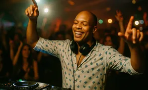 A bald Black man, wearing a patterned shirt and headphones, smiles and points up while DJing at a club.