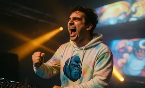 A man in a tie-dye hoodie with a gorilla print shouts while raising his fist, illuminated by yellow stage lights.