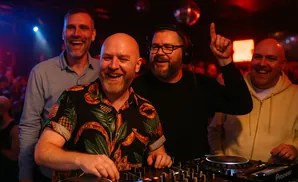 Four men in a club setting, two behind DJ equipment, all smiling and looking happy.