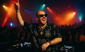 DJ with arm raised in a rock-on gesture, wearing sunglasses and a patterned shirt, performing at a club with colorful lights.