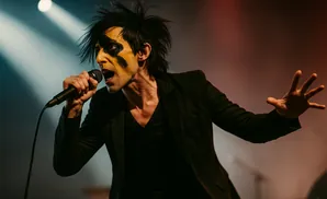 A man with theatrical makeup sings into a microphone on stage.