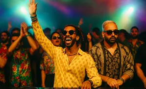 DJs in a club with colorful lights, one with his arm raised, the other looking forward.