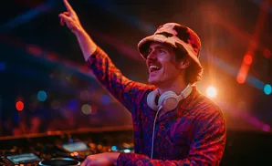 A DJ in a patterned shirt and bucket hat smiles, pointing a finger up while working at a DJ booth with colorful lights.