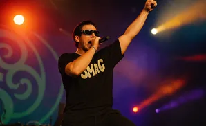 A rapper in sunglasses performs on stage, holding a microphone and raising his fist in the air under a spotlight.