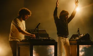 Two musicians on stage at night, one with hands raised, the other at a DJ console.