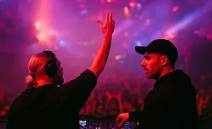 Two DJs in a club, one with headphones raising his arm, the other in a cap operating equipment.