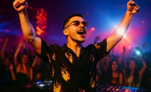 A DJ in a flame shirt with arms raised in celebration, with a crowd in the background under colorful lights.