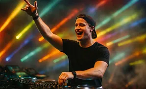 A smiling DJ in a black shirt and cap, with his arm raised, plays music at a console against a backdrop of colorful lights.