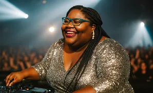 A joyful Black woman in a sequined dress and teal glasses laughs while DJing at a concert with spotlights and a blurred crowd.