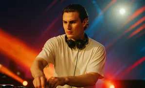 A young man with headphones around his neck works a DJ mixer, illuminated by vibrant orange and blue stage lights.