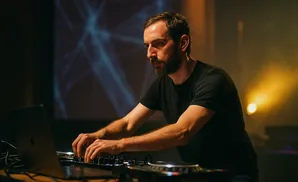 Man with a beard in a black t-shirt operating DJ equipment and a laptop with a blue light display behind him.
