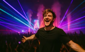 A man with red hair laughs with his arms outstretched on a stage with purple and blue laser lights and a cheering crowd.