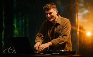 A smiling DJ works on his laptop and equipment with warm stage lighting.