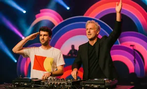 Two DJs perform with a keyboard and turntables, bathed in warm stage lighting.