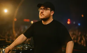A man wearing a black t-shirt and black baseball cap DJing in front of a crowd with lights.