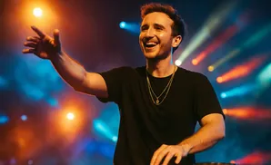 A smiling man in a black t-shirt DJs on a stage with colorful lights.
