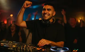 A DJ in a black t-shirt with headphones around his neck raises his fist in celebration while working at a DJ booth in a dimly lit club.