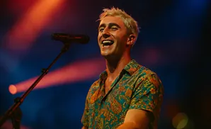 A man with blonde hair sings into a microphone on a stage, wearing a colorful patterned shirt.