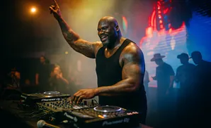 DJ Shaquille O'Neal in a black tank top spins records with his arm raised in a club with colorful lighting.