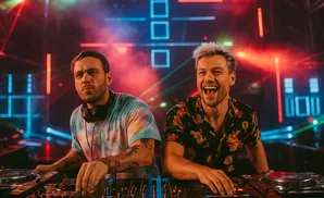 Two DJs at a console with colorful lights and smoke in the background.