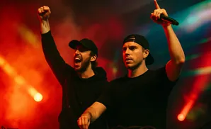 Two DJs in black t-shirts and baseball caps on stage with red and blue lights.
