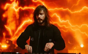 DJ with long brown hair and a beard, wearing a black jacket, plays music on a DJ console against a vibrant orange, lightning-like background.