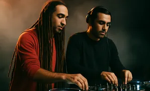 Two DJs with dreadlocks and headphones mix music on turntables in a dimly lit studio.