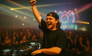 DJ with arm raised in celebration at a lively concert with colorful stage lights and a cheering crowd.