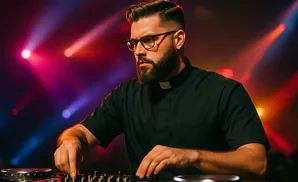 A man with glasses and a beard wearing a priest's collar works a DJ mixer, with colorful stage lights in the background.