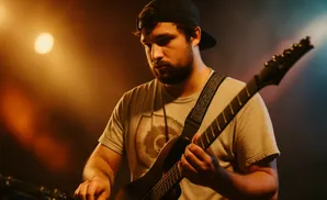 Man in a black baseball cap playing an electric guitar on stage under warm lighting.