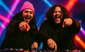 Two DJs in black hoodies and a pink beanie laugh and point at the camera, with colorful stage lights in the background.