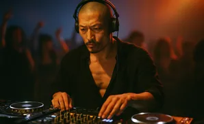 A bald DJ wearing headphones, intently mixing music on a console in a dimly lit club with a crowd in the background.
