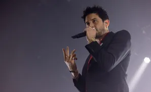 Man singing into a microphone on stage, wearing a suit jacket and jewelry.