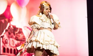 Singer Melanie Martinez in a white ruffled dress, holding a microphone on stage with a pink and balloon background.