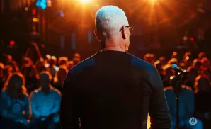 Man with white hair and glasses on stage addresses an audience in a dimly lit venue with warm lighting.