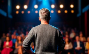 a rear-view photograph from behind american television host and comedian greg gutfeld, delivering a live stand-up monologue on stage in front of a large audience.