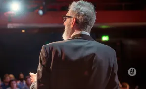 Rear view of a man with gray hair and beard in a suit, speaking to an audience.
