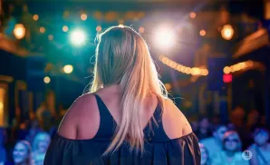 Back view of a woman with blonde hair on stage, facing bright lights and an audience.