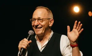 Man in glasses and vest smiles while holding a microphone and raising his hand.