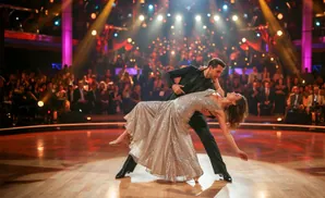 A couple dances ballroom on a shiny wooden floor, lit by stage lights, with an audience in the background.