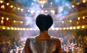 Woman in a sparkly dress stands on stage in a packed, ornate theater.