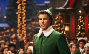 Will Ferrell as Buddy the Elf in a green elf costume with a white fur collar, standing in front of Christmas trees and lights.
