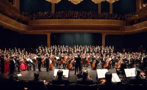 An orchestra and choir perform in a grand concert hall.