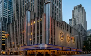Art Deco buildings in NYC, featuring ornate mosaic medallions and illuminated blue trim, are seen at dusk.