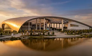 A modern stadium with a curved, metallic roof reflects in a body of water at sunset.