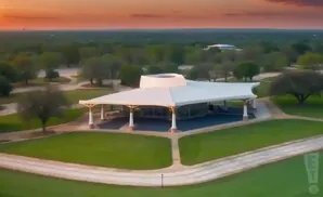 An aerial view of a large, white, open-air pavilion with a unique roof design on a green lawn during sunset.