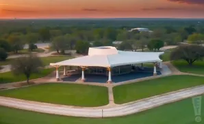 An aerial view of a white pavilion with a unique roof on a grassy area at sunset.